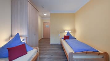 A room with two single beds, each with a blue mattress and a red pillow. There is a wooden wardrobe on one side and a door at the end of the room.