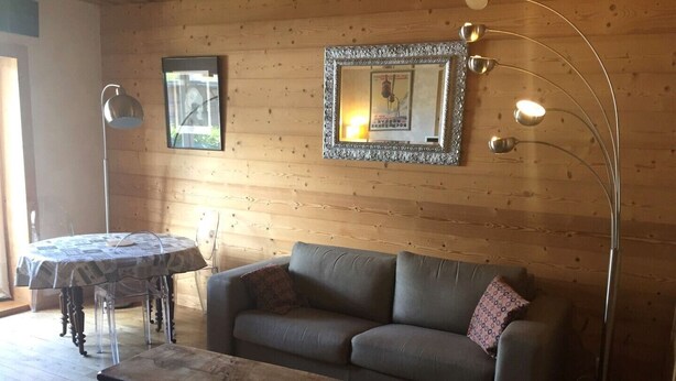 A cozy living room with a wooden wall, a grey sofa, a wooden coffee table, a round table with a patterned tablecloth, and a floor lamp. - MEGEVE-T2-44M²-NEAR TOWN CENTER (Megève)