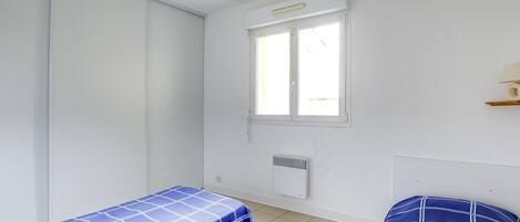 A small, clean room with two single beds, a window, a wall-mounted heater, and a hanging light fixture.