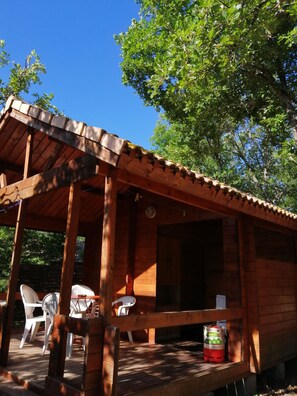 A wooden cabin with a covered porch, white plastic chairs, and a propane tank on the deck. - Chalet Acanthe (Simiane-la-Rotonde)