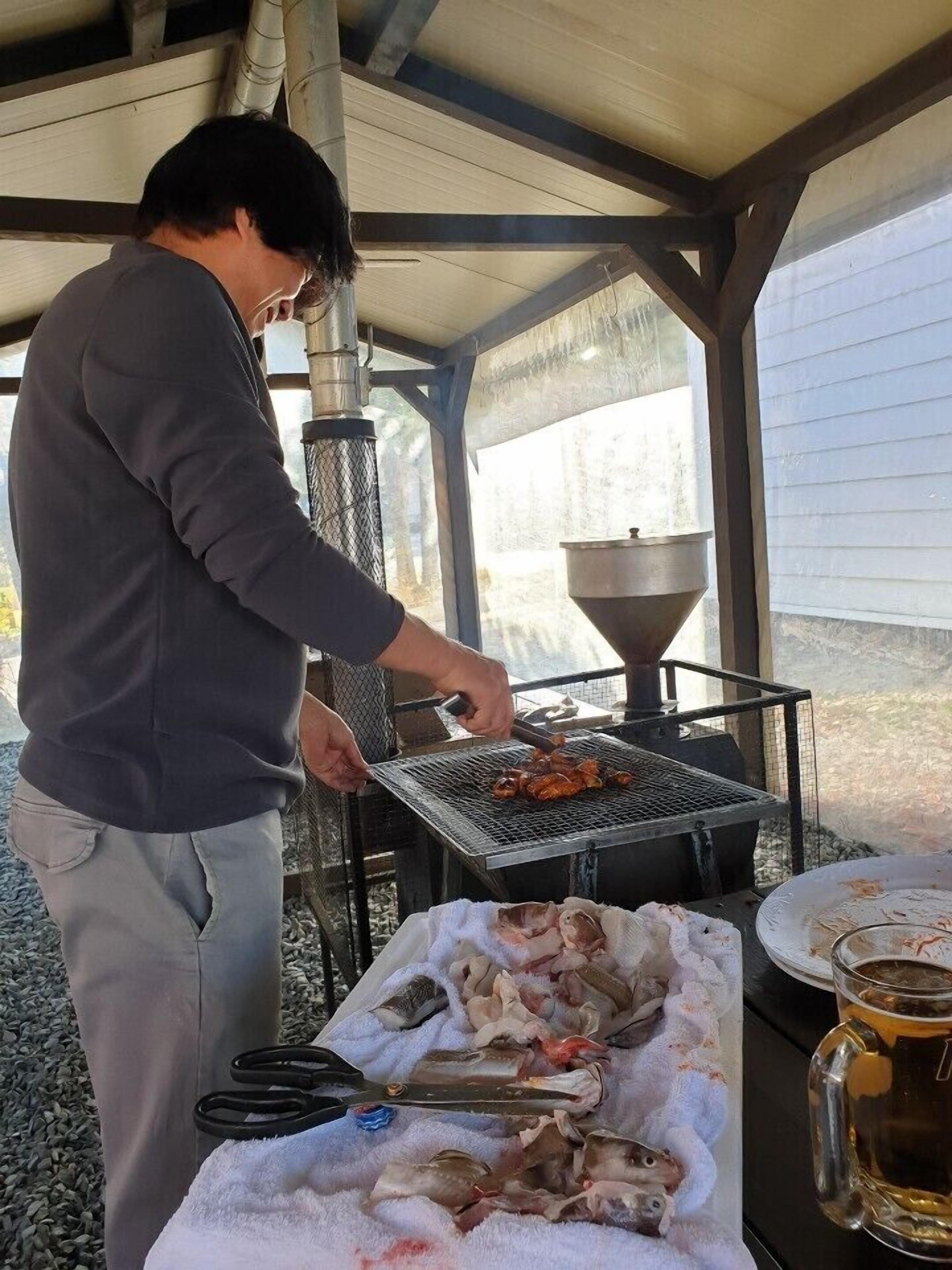 A person grilling seafood outdoors with a grill and a table with a bowl of fish and a beer mug.