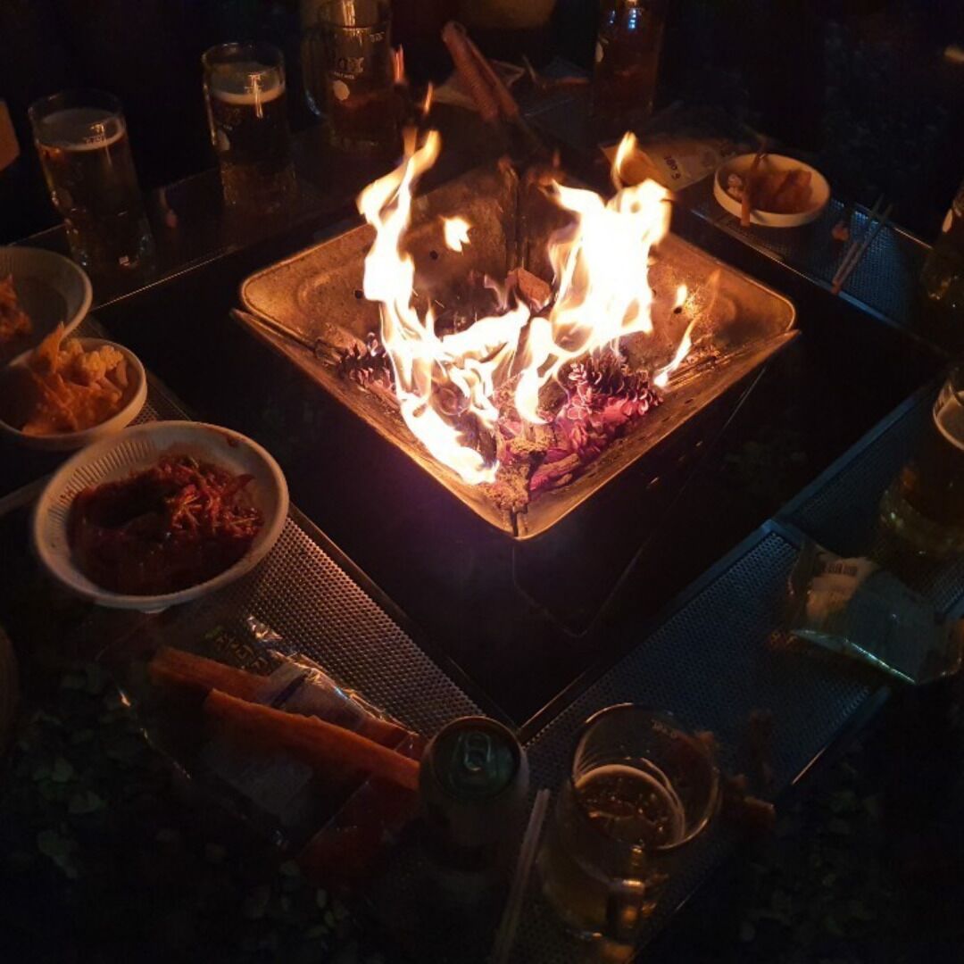 Ein Tisch mit einem zentralen Feuer, umgeben von verschiedenen Speisen und Getränken, was auf eine Esssituation hindeutet.