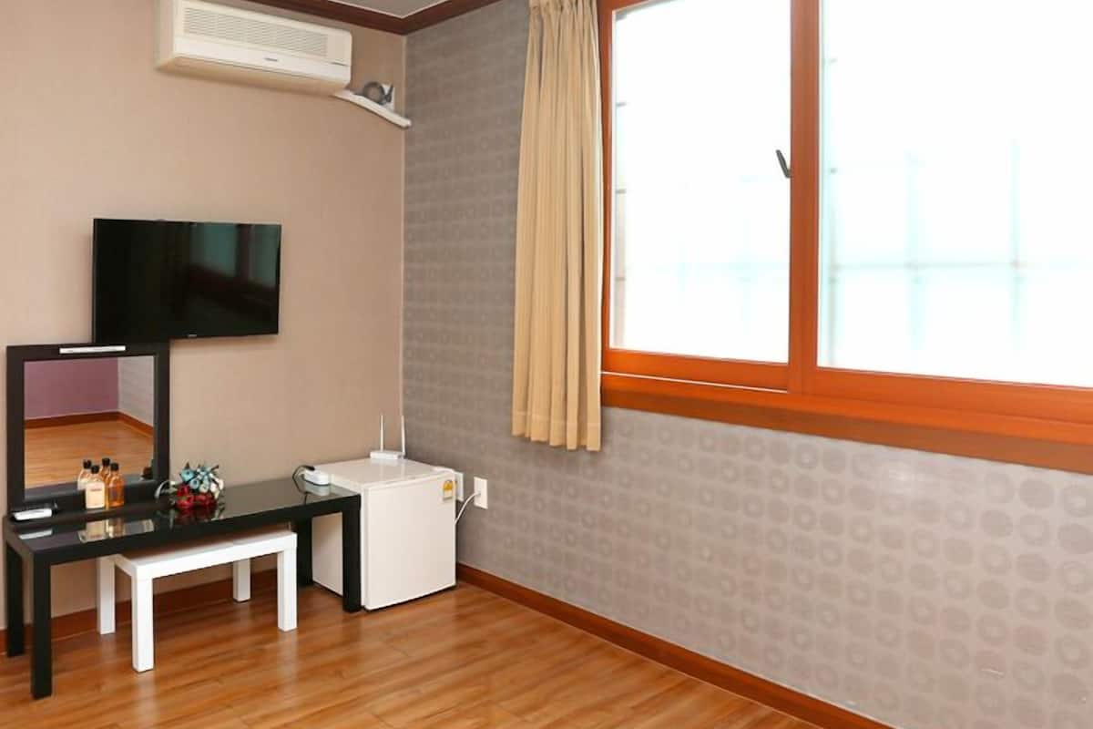 A hotel room with a wooden floor, a flat-screen TV mounted on the wall, a small white desk with a mirror, a white radiator, and a large window with a curtain.