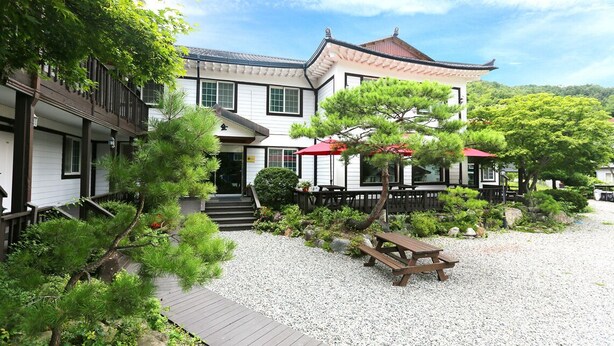 A traditional Japanese-style building with a wooden deck, outdoor seating, and a well-maintained garden with pine trees and gravel pathways.