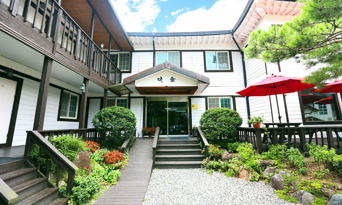 A traditional Japanese building with a wooden staircase leading to a glass entrance, surrounded by a garden with rocks and plants.