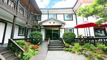 A traditional Japanese building with a wooden staircase leading to a glass entrance, surrounded by a garden with rocks and plants.