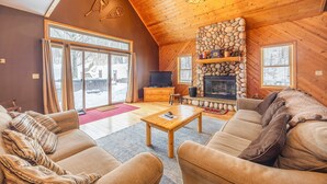 A cozy living room with a stone fireplace, wooden ceiling, and a large window overlooking a snowy landscape.