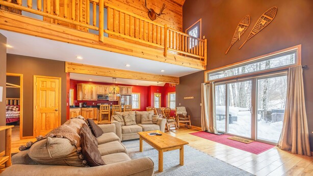 A spacious living room with a high wooden ceiling, a cozy seating area, and a large sliding glass door leading to a snowy outdoor area.