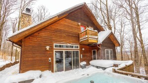 A wooden cabin with a hot tub surrounded by snow, a stone chimney, and a balcony.