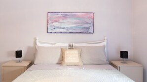 Quarto bem arrumado com uma cama, duas mesas de cabeceira, um quadro abstrato na parede e um travesseiro com uma frase motivacional.