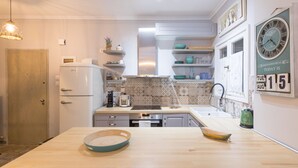 Cozinha moderna com armários brancos, bancada de madeira e forno embutido. Há um relógio na parede e uma prateleira com louças.