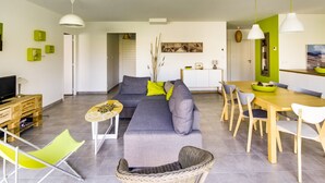 A modern living room with a sofa, dining table, and chairs. There is a television and a small table with a lamp. - Modern Home with Terrace in Biscarrosse – Sleeps 7 with WiFi & BBQ (Biscarrosse)