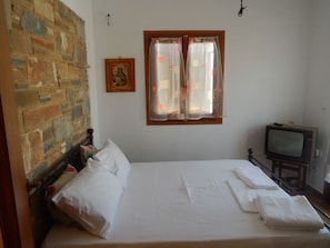 A room with a bed, a stone wall, a window with curtains, and a television. - Armonia Studios and Villas (Naxos)