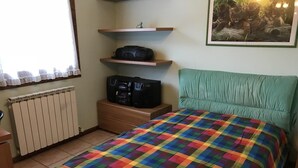 A bedroom with a colorful checkered bedspread, a radiator, a shelf with electronic devices, and a framed picture on the wall. - Apartment with garden and parking (Edolo)