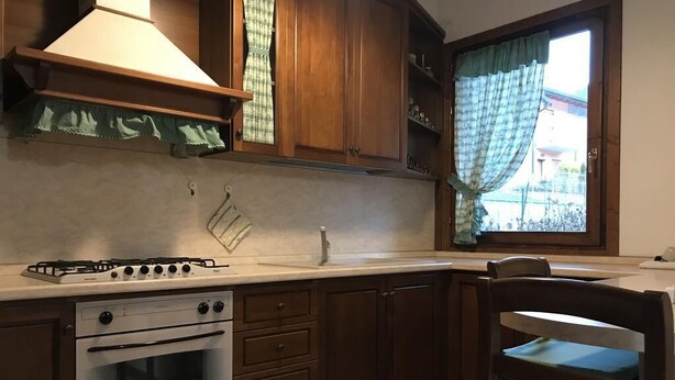 A kitchen with wooden cabinets, a gas stove, a built-in oven, and a window with curtains. - Apartment with garden and parking (Edolo)