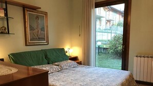 A bedroom with a bed, a bedside table, a lamp, a framed picture on the wall, and a view of the outside. - Apartment with garden and parking (Edolo)