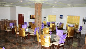 Uma sala de jantar com mesas, cadeiras, um pilar e uma televisão na parede.