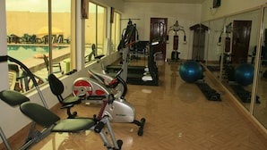 Ginásio com equipamentos de exercício, vista para a piscina e parede com espelhos.