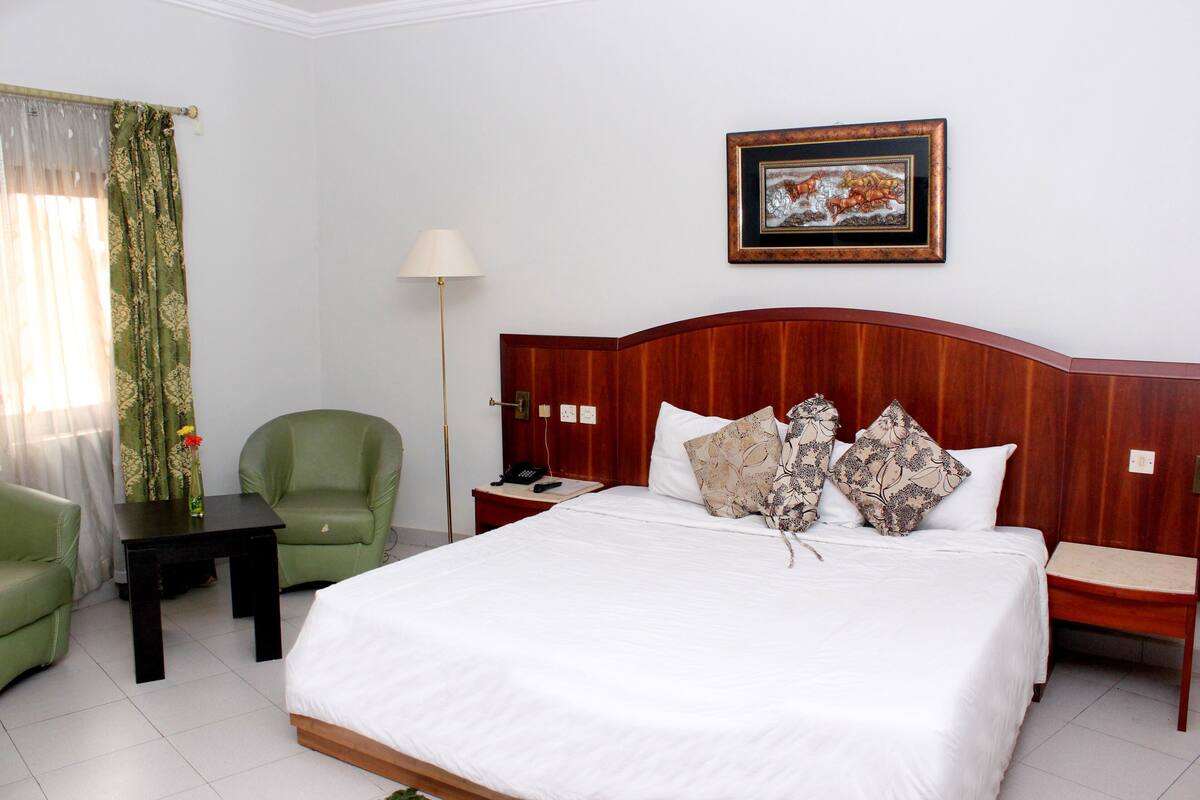 A hotel room with a bed, a green armchair, a nightstand, a lamp, and a framed picture on the wall.