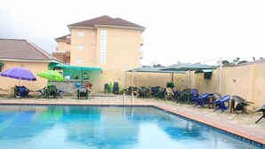 Uma piscina com espreguiçadeiras e guarda-sóis, cercada por um muro baixo e um edifício ao fundo.