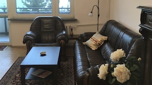 En stue med en mørk lædersofa, en matchende lænestol, et sofabord og et stort vindue med udsigt til bygninger og træer.