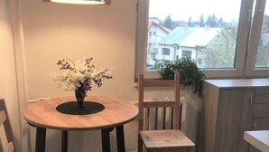 Et lille køkken med et rundt træbord, to stole, en hængende pendellampe, et vindue med udsigt til bygninger og en potteplante på vindueskarmen.