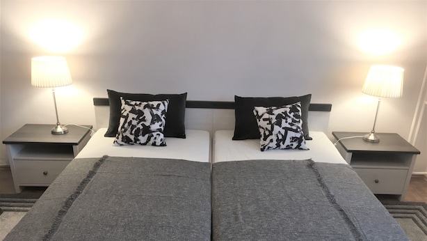 En dobbeltseng med et gråt tæppe, to puder med sort-hvidt mønster og to sengetårnslamper på matchende sengeborde.