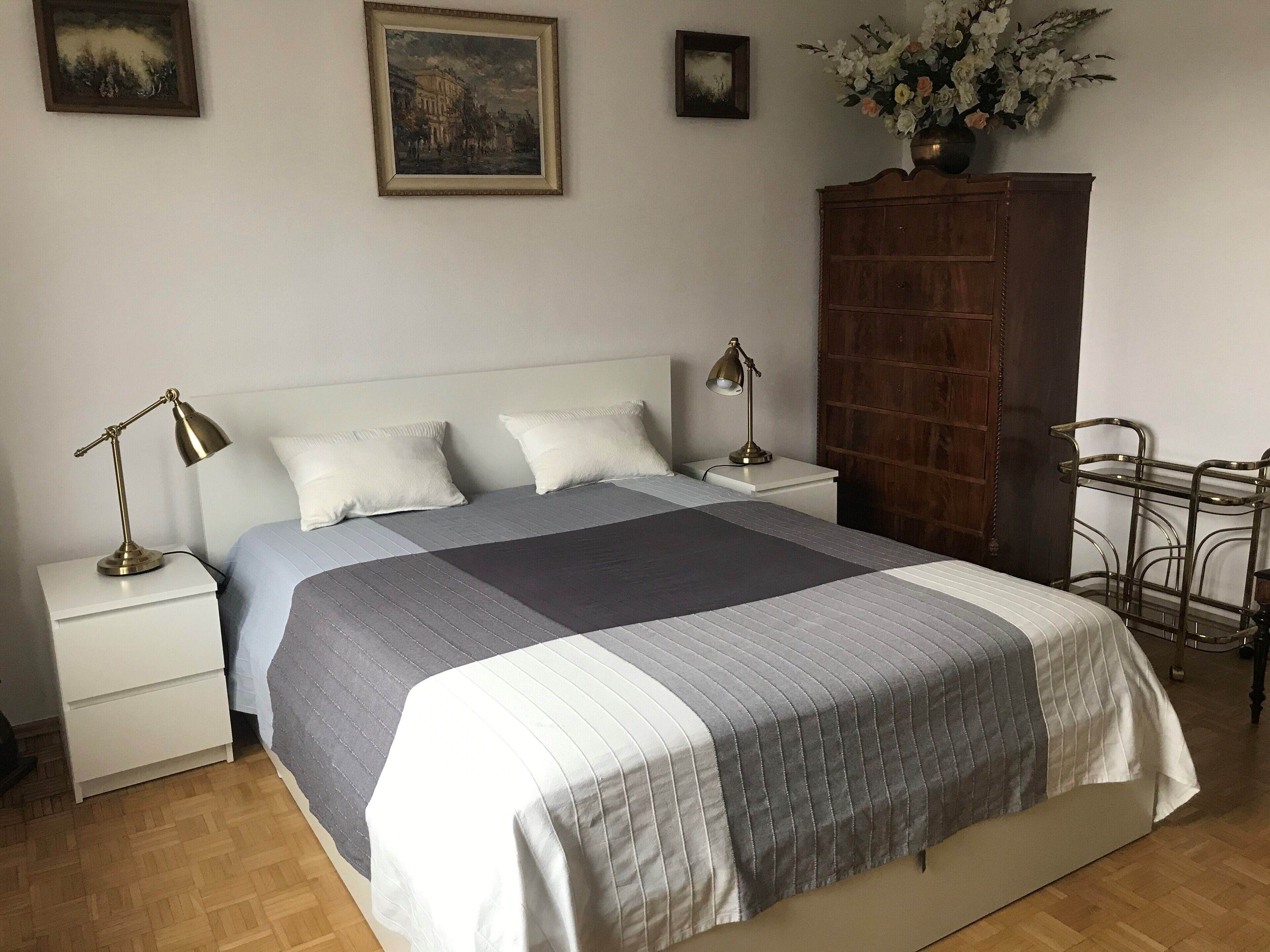 Una cama bien hecha con una colcha a rayas, dos lámparas de noche, una mesita de noche, un armario de madera y un perchero metálico.