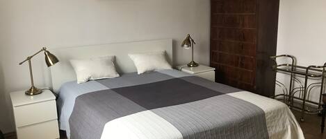 Una cama bien hecha con una colcha a rayas, dos lámparas de noche, una mesita de noche, un armario de madera y un perchero metálico.