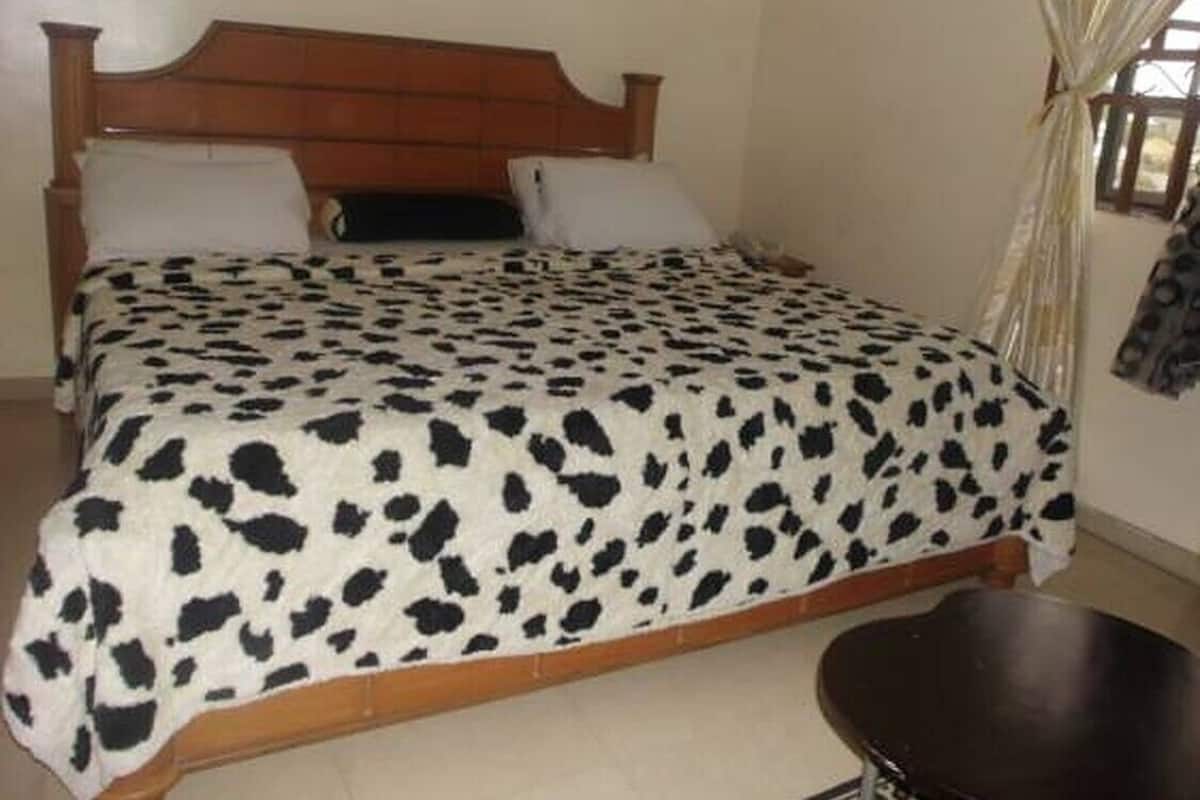 Un lit avec une couvre-lit à motifs, une tête de lit en bois et une chaise dans une pièce aux murs unis et dotée d’une fenêtre avec des rideaux.