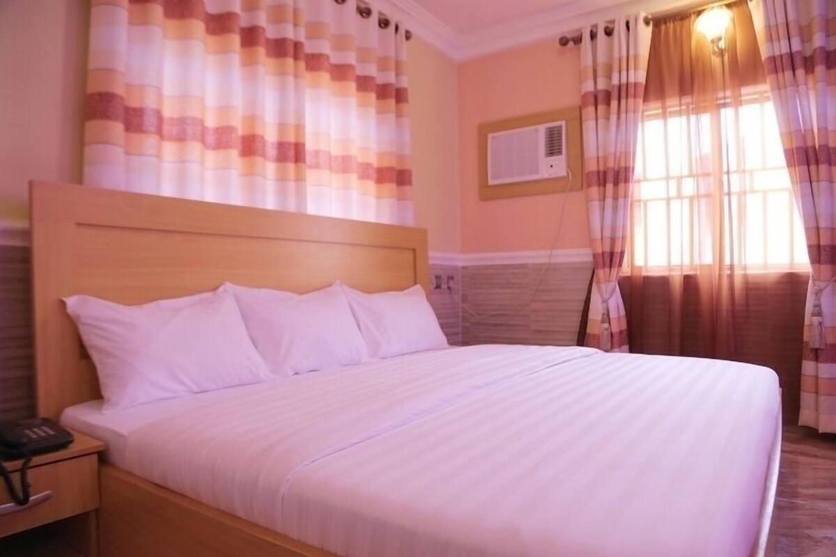 A hotel room with a large bed, a wooden headboard, a nightstand with a phone, and a window with curtains.