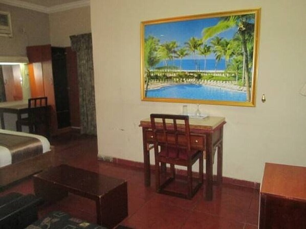 A hotel room with a bed, a desk with a chair, a painting of a beach scene, and a small table.