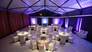 Un salón de banquetes con mesas redondas, sillas blancas y un escenario.