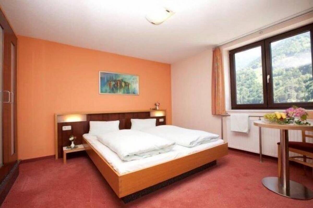 Hotellihuone, jossa on sänky, yöpöytä, työpöytä tuolilla, ikkuna, josta avautuu näkymä vehreään maisemaan, ja taulu seinällä.