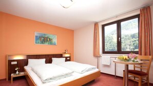 A hotel room with a double bed, a small table with a chair, a painting on the wall, and a window with curtains. - Gasthof Stern (Vandans)