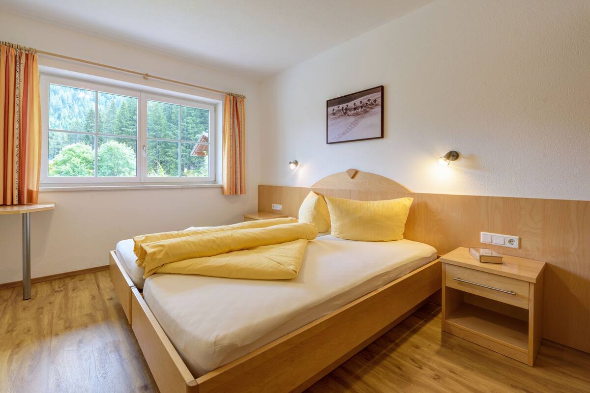 A neatly made bed with a wooden headboard, a bedside table with a lamp, and a window with curtains.