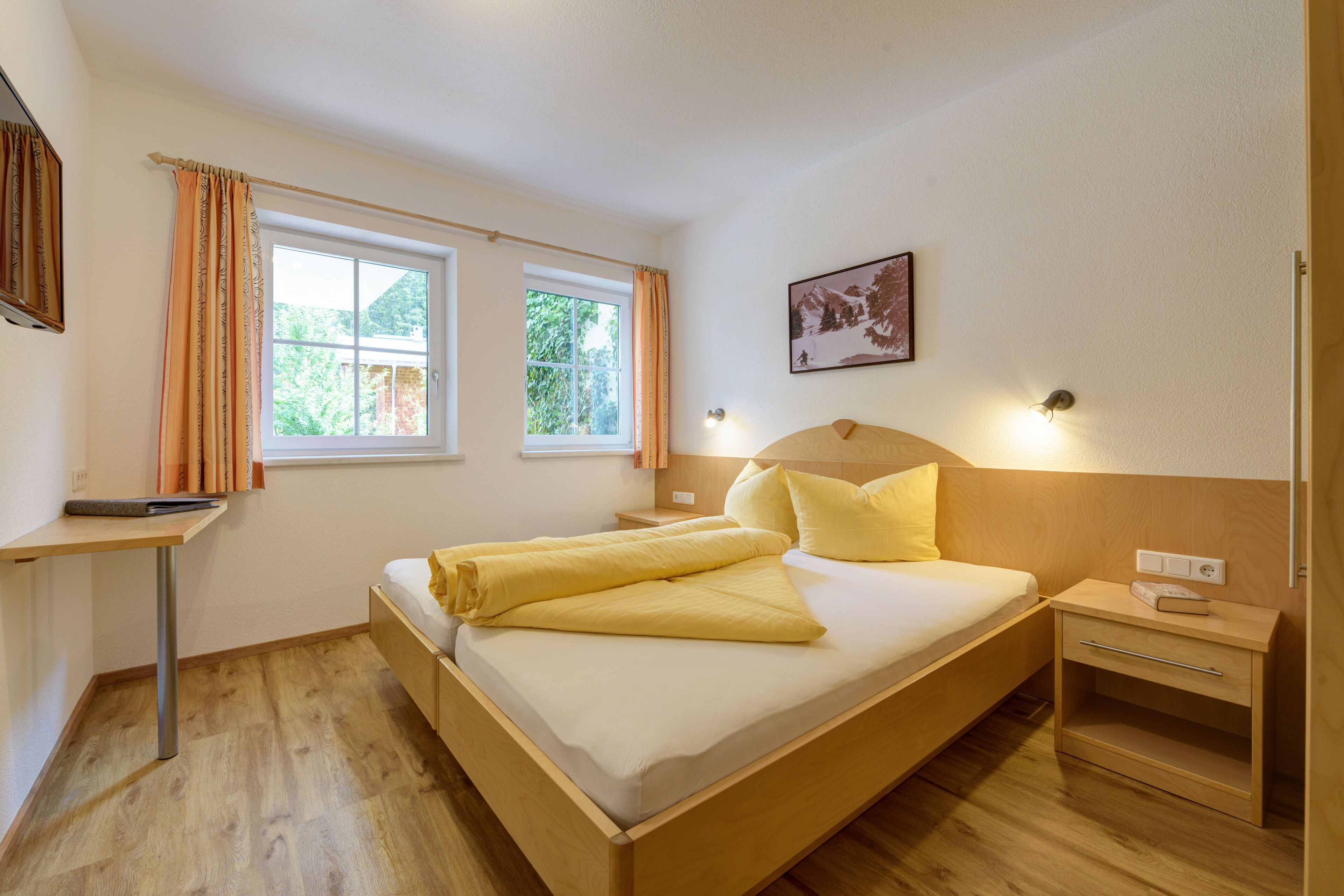 Un dormitorio ordenado con cama, mesitas de noche, escritorio y ventana con cortinas.