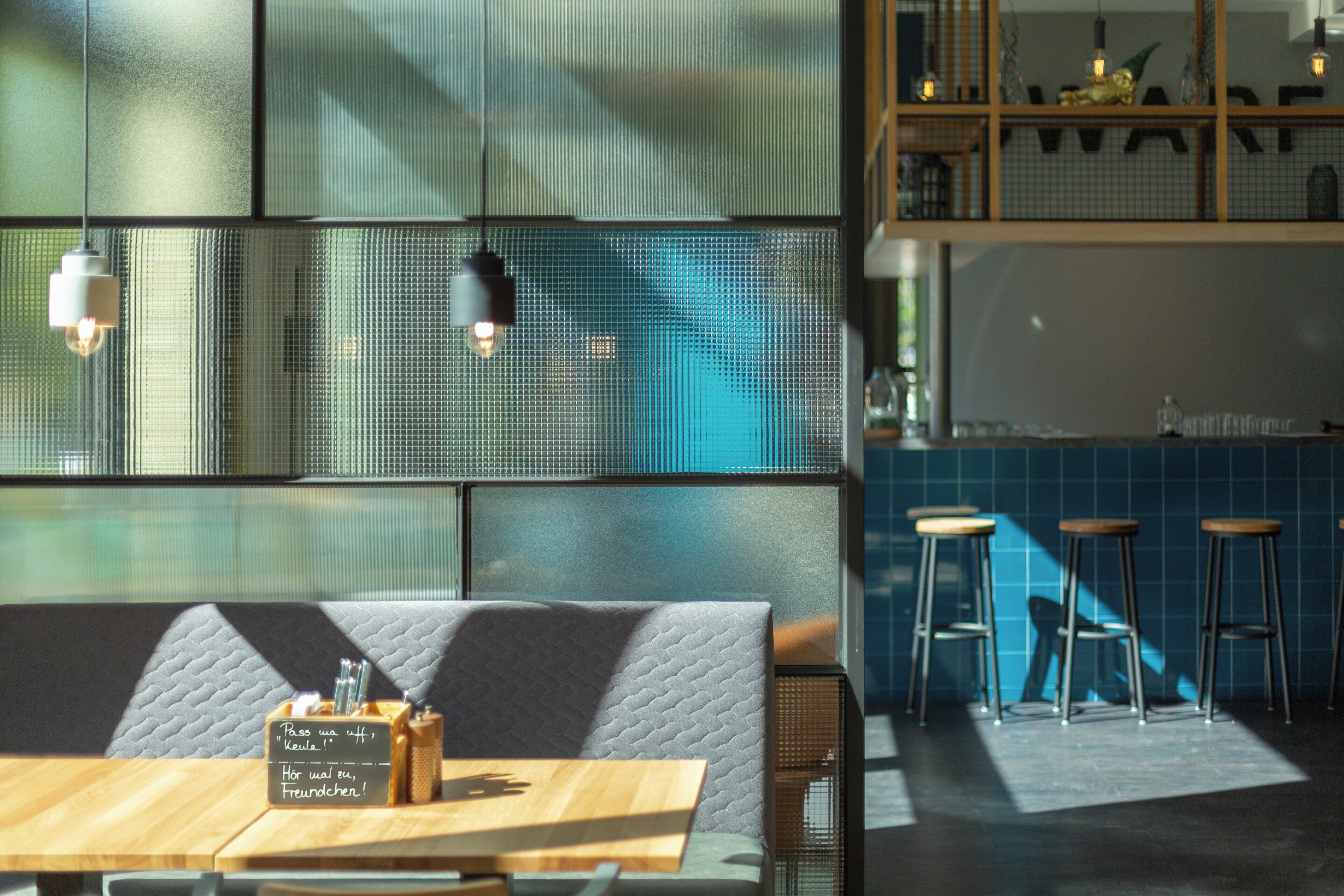 Een restaurantinterieur met een houten tafel, zachte zitplaatsen, hangende lampen en een blauwe betegelde achterwand.