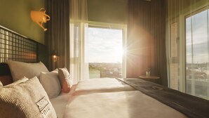 A hotel room with a large bed, a view of the city through the window, and a small table with a lamp. - Holiday Inn - the niu, Dwarf Berlin Schöneberg by IHG (Berlin)