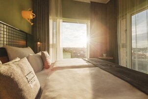 Ein Hotelzimmer mit einem großen Bett, Blick auf die Stadt durch das Fenster und einem kleinen Tisch mit einer Lampe.