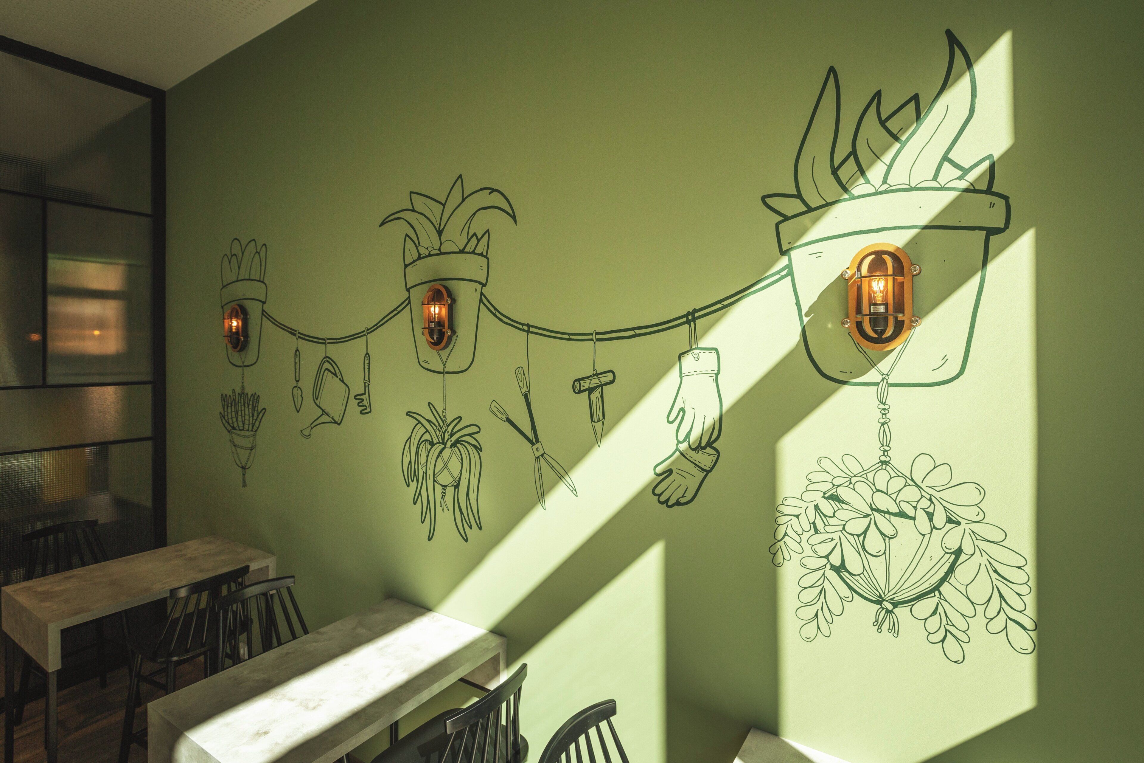 A dining area with tables and chairs, a wall with hanging light fixtures, and a green wall with plant illustrations.