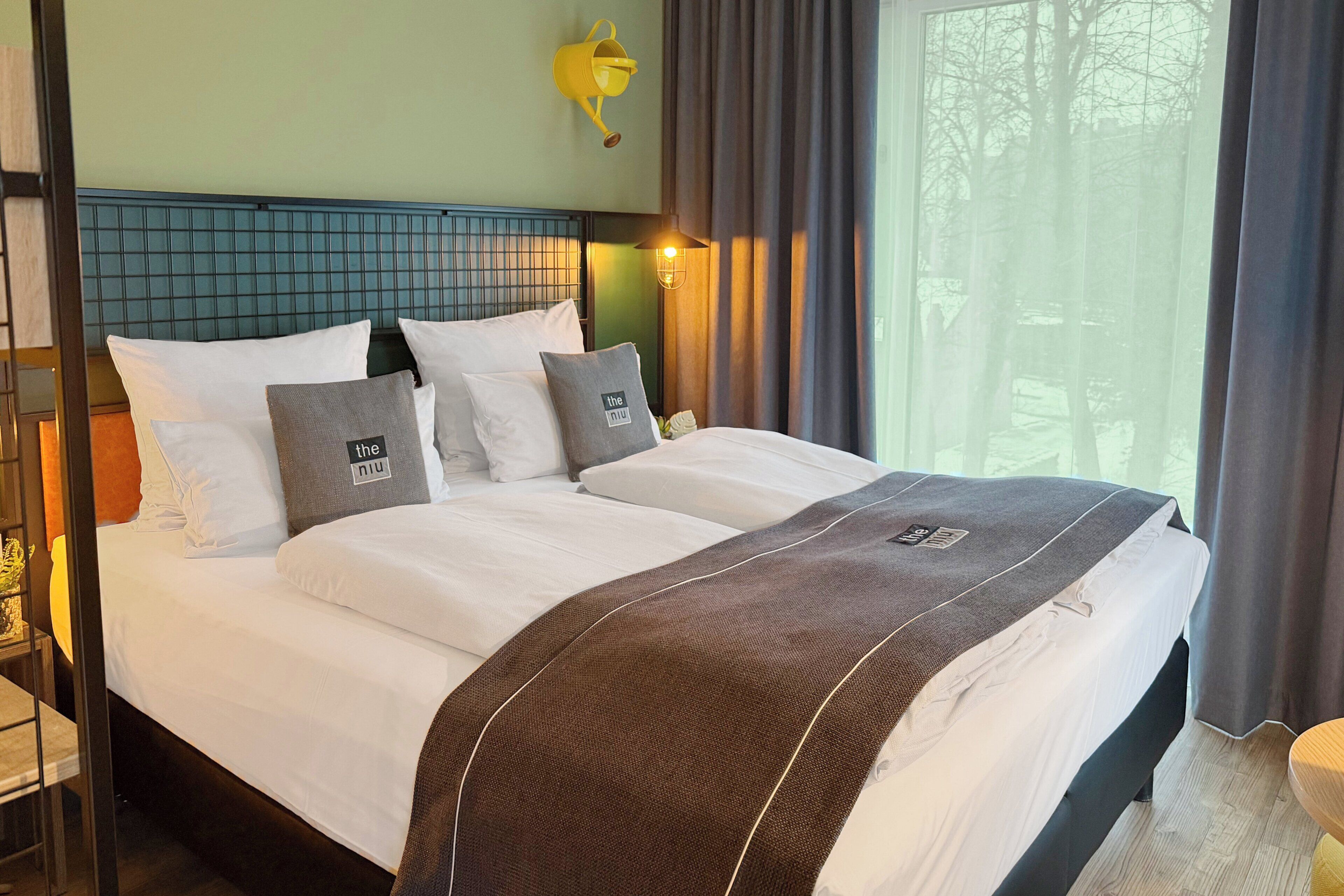 A hotel room with a bed, a nightstand, a lamp, a window with curtains, and a view of trees.