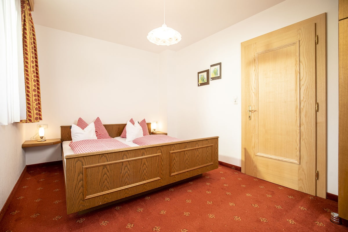 A bedroom with a wooden bed, a red carpet, a wooden door, and a white pendant light.