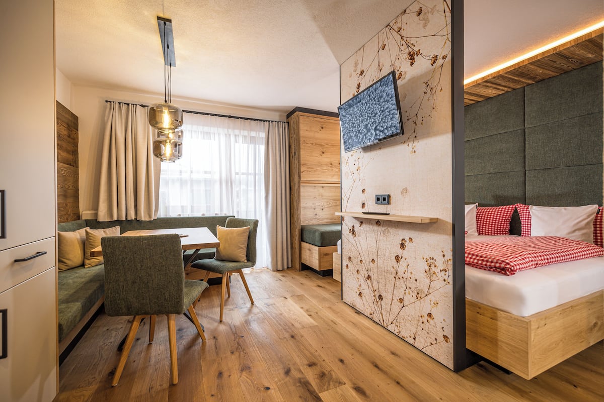 A modern room with a wooden floor, a sofa, a dining table with chairs, a bed with red checkered bedding, and a mounted TV.