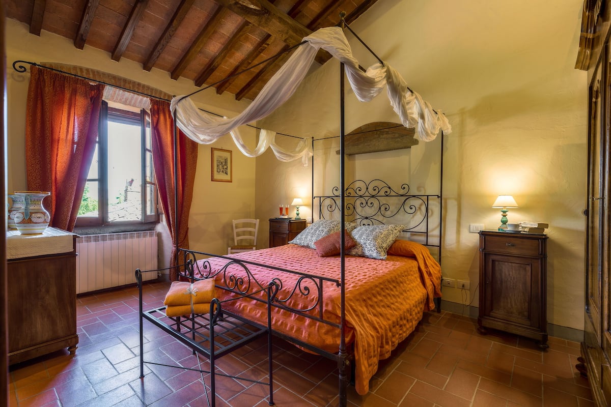 A bedroom with a four-poster bed, a wooden floor, a window with curtains, a bedside table with a lamp, and a radiator.