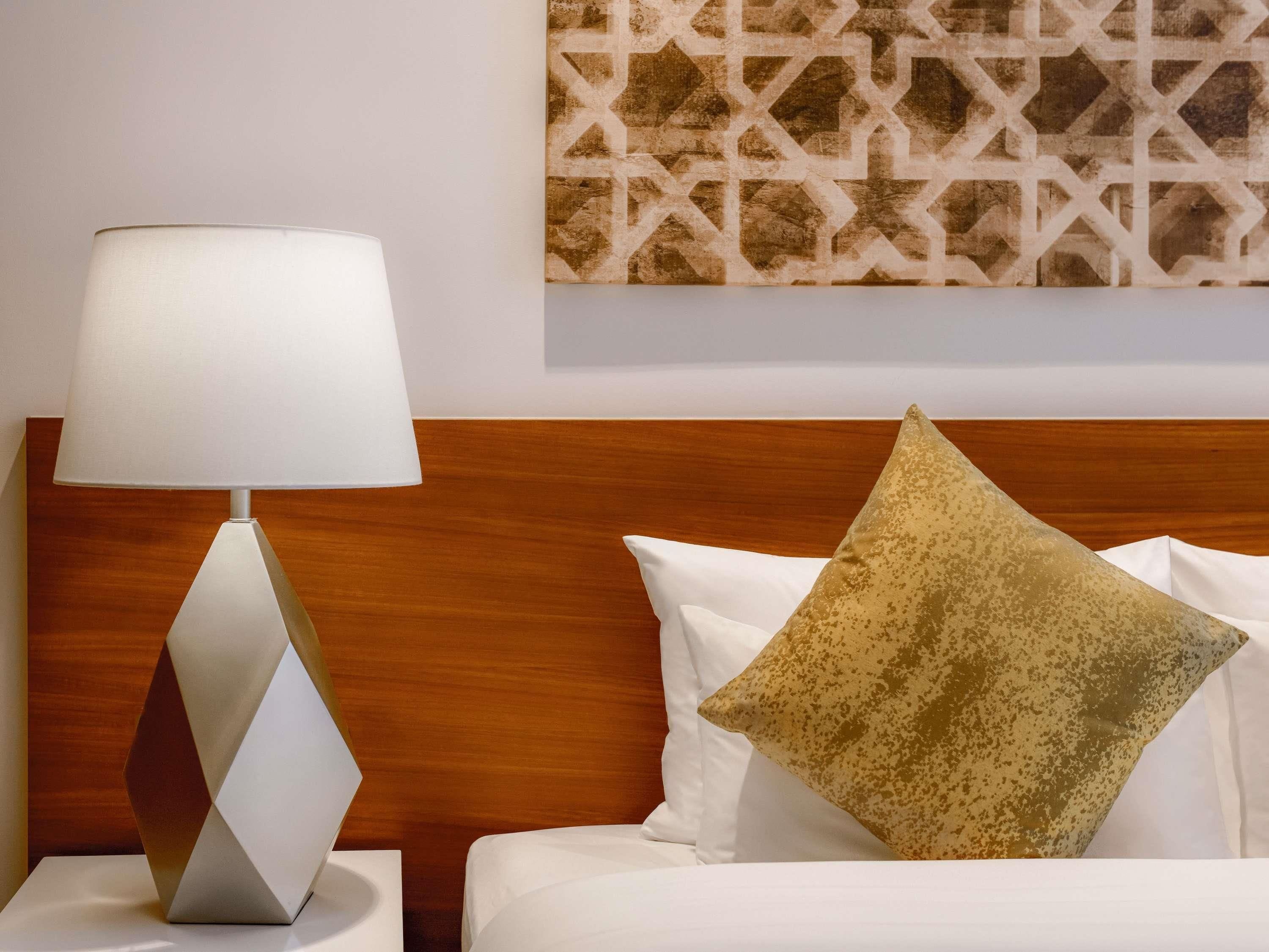 A bed with white bedding and a single gold-patterned pillow, a modern lamp with a white shade, and a geometric wall art piece.