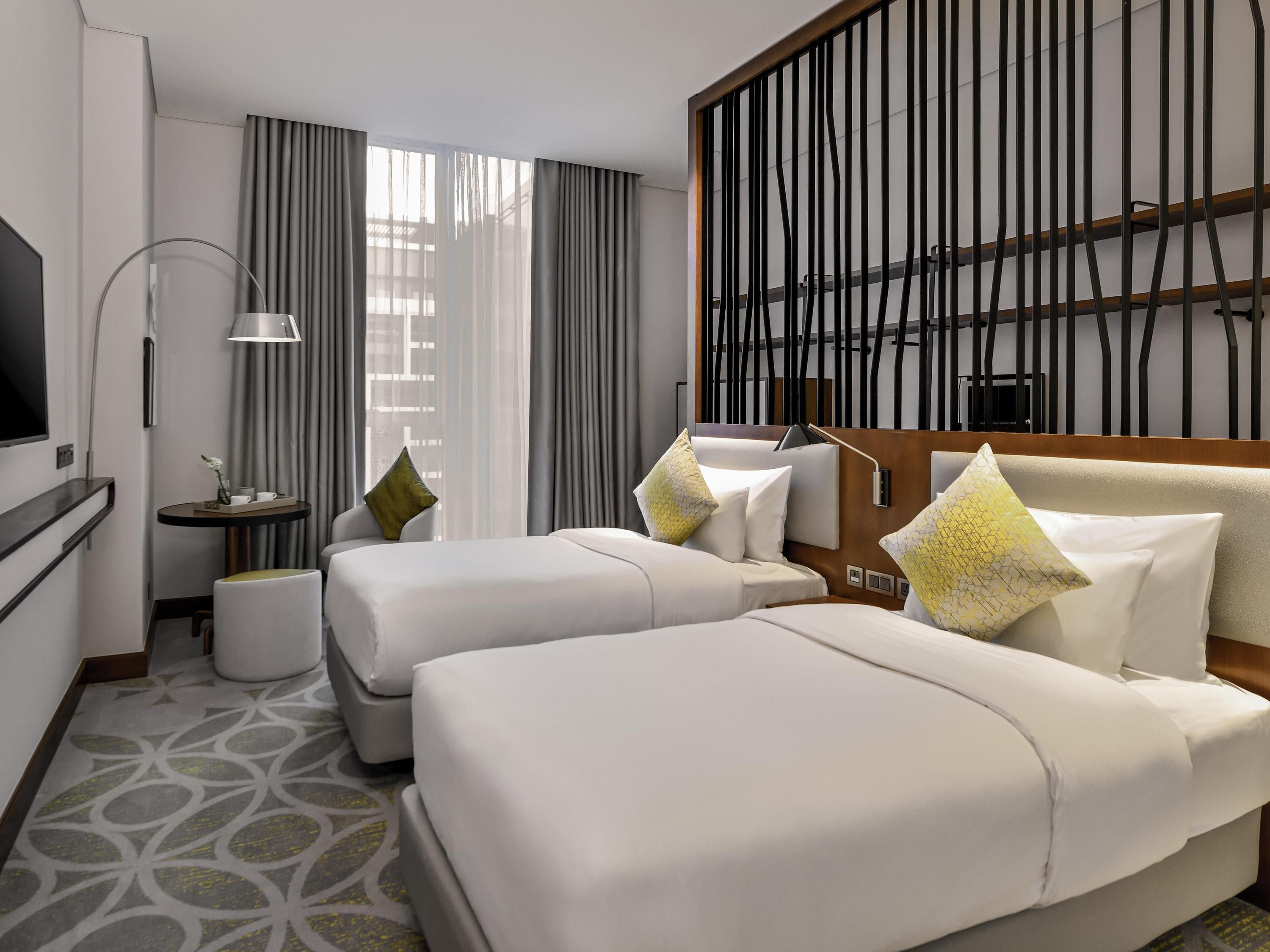 A hotel room with two beds, a TV, a desk, and a large window with curtains.