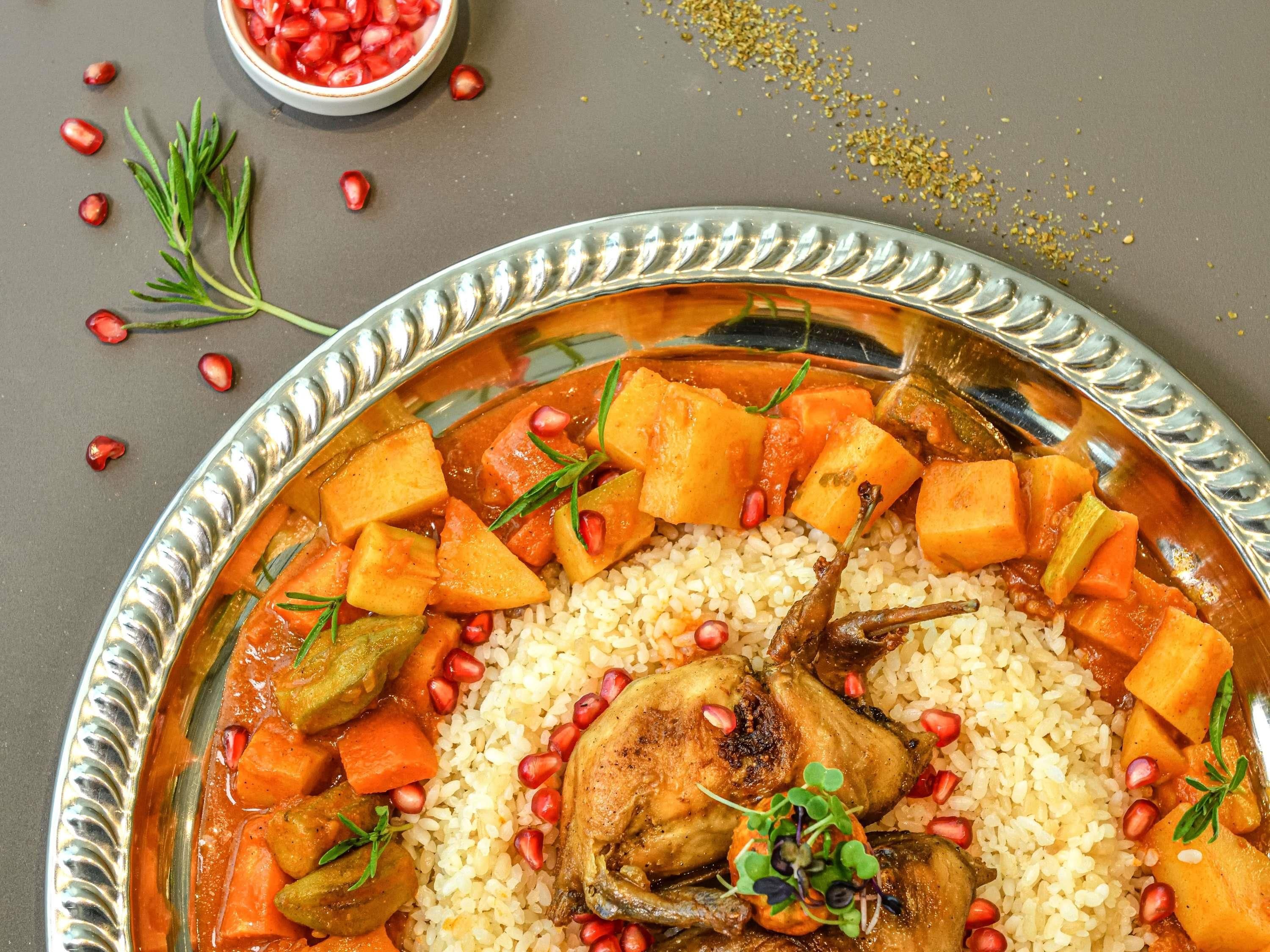 Una gran bandeja de plata ornamentada llena de arroz, pollo asado y un guiso de verduras y semillas de granada.