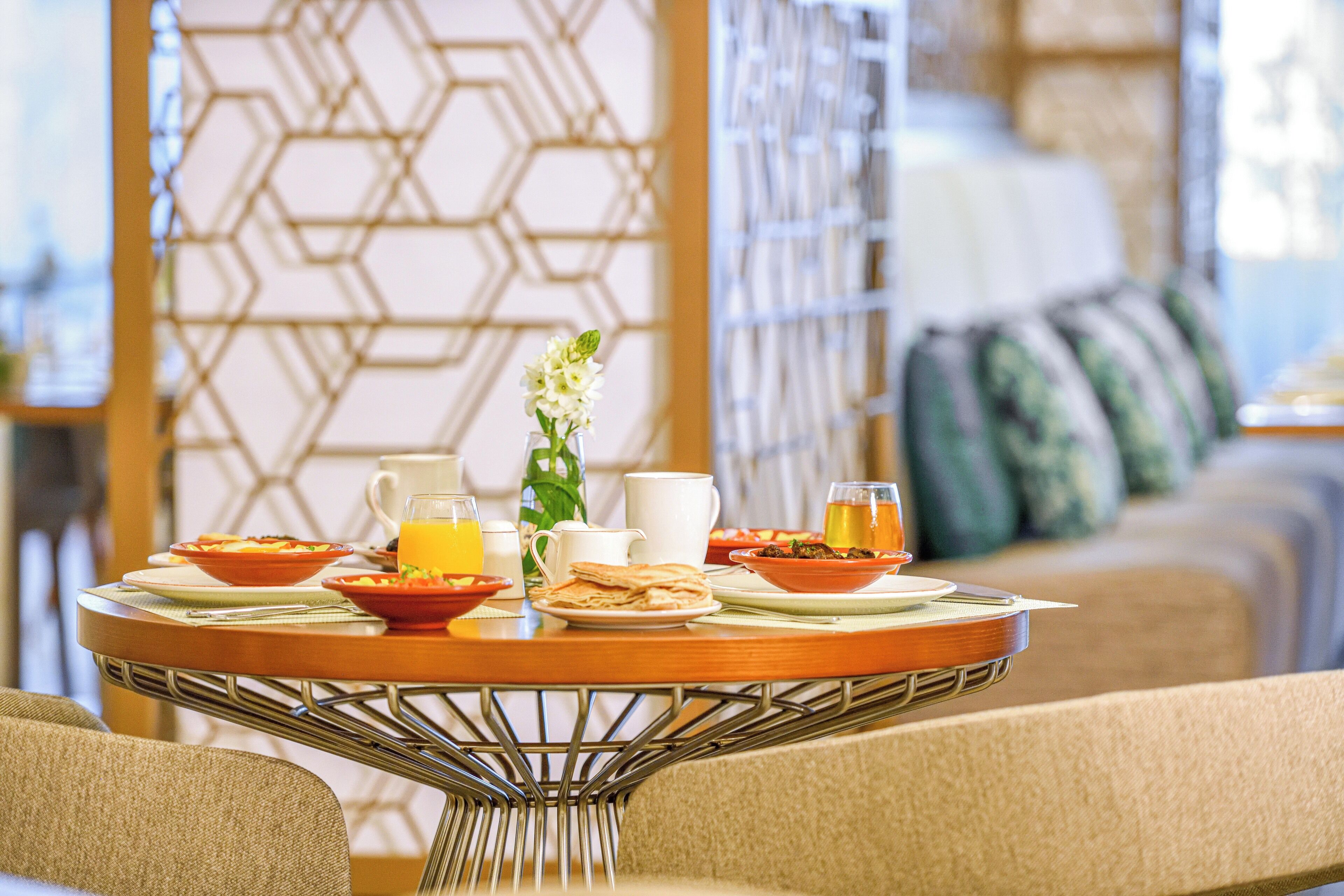 A breakfast table with various dishes and drinks, a vase with a flower, and a patterned wall in the background.