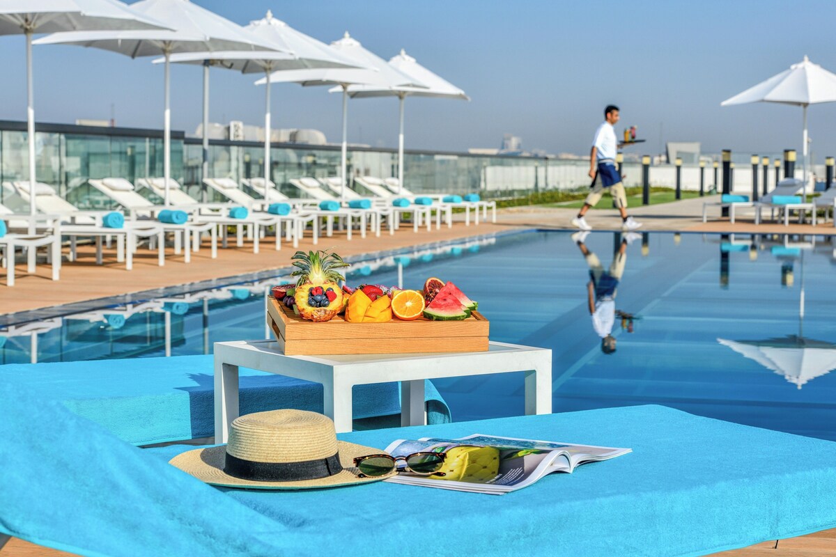 Un espace aménagé au bord de la piscine, avec des chaises longues, des parasols et une table sur laquelle sont disposés des fruits et un chapeau.